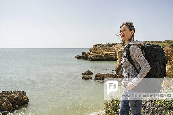 Glückliche Frau mit Rucksack  die an einem sonnigen Tag vor dem Meer steht