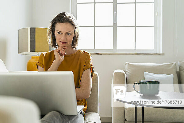 Smiling freelancer with hand on chin looking at laptop siting in living room
