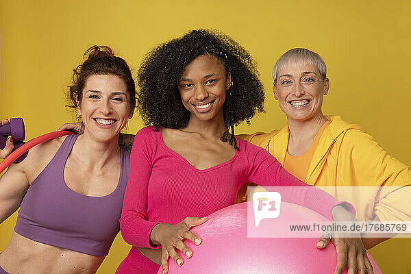 Smiling multiracial friends with fitness equipment standing against yellow background