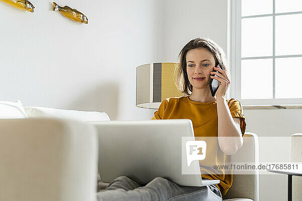 Smiling freelancer talking on mobile phone sitting with laptop on sofa in living room
