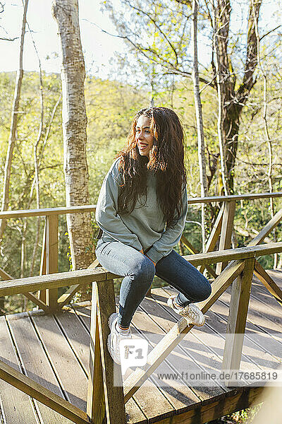 Smiling young woman sitting on wooden railing of bridge