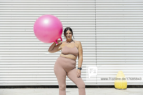 Curvy beautiful woman holding pink fitness ball standing in front of corrugated wall