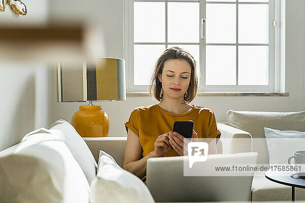 Freelancer using smart phone sitting with laptop on sofa at home