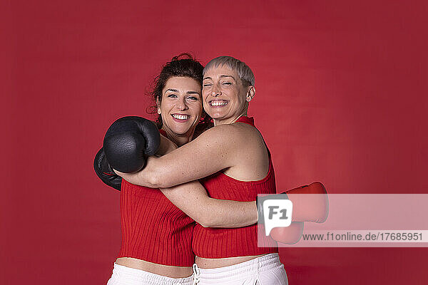 Smiling friends wearing boxing gloves hugging each other against red background