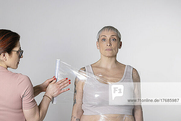 Woman wrapping friend in plastic foil against white background