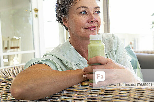 Smiling mature woman with bottle of smoothie sitting on sofa at home