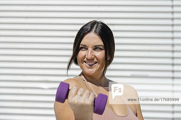 Happy curvy woman exercising with dumbbell exercising in front of corrugated wall
