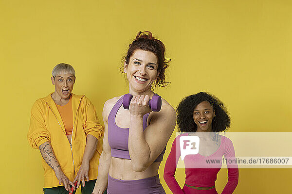 Smiling woman holding dumbbell standing with friends against yellow background