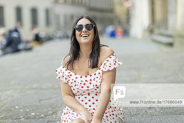 Glückliche Frau mit herzförmiger Sonnenbrille sitzt auf Fußweg