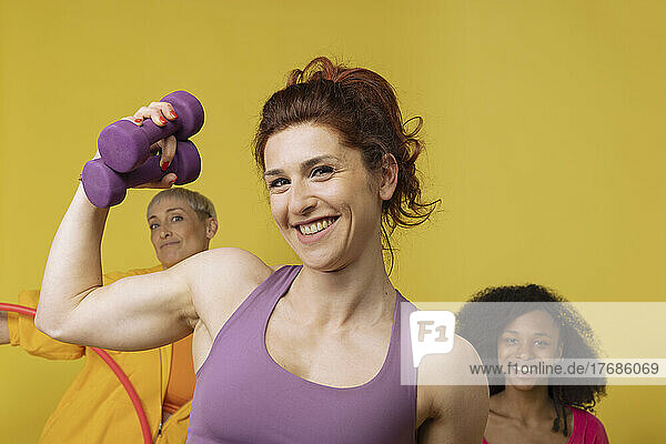 Happy woman holding dumbbell standing with friends against yellow background