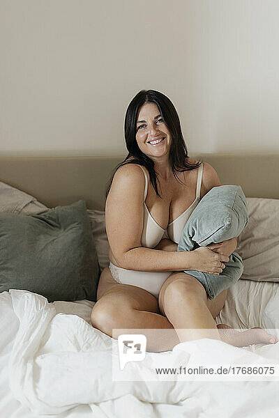 Smiling plus size woman holding pillow sitting on bed at home