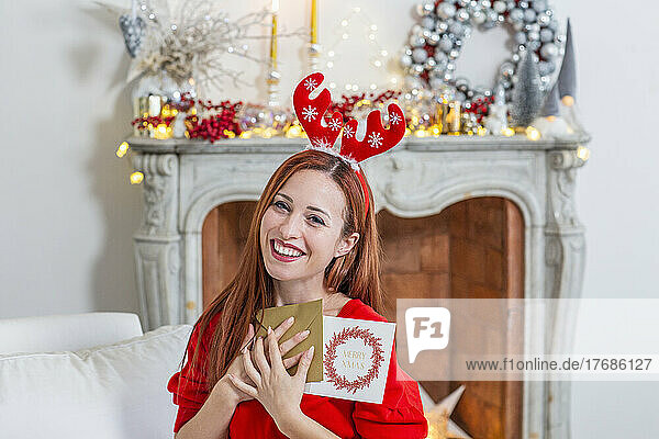 Happy woman wearing Christmas headdress holding greeting cards at home