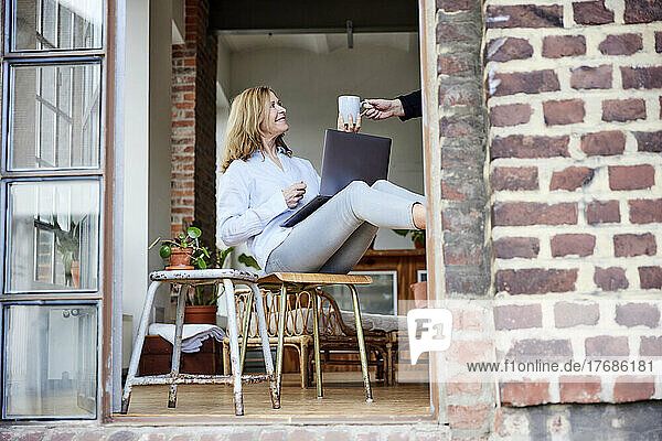 Smiling freelancer sitting on chair with laptop taking coffee cup