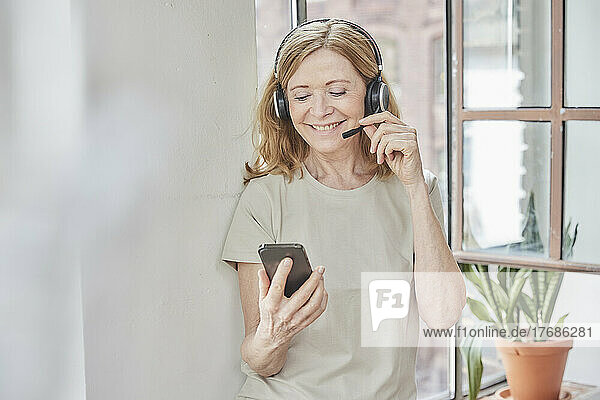 Smiling woman with headset using smart phone standing in front of window at home