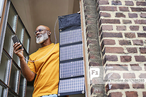 Man with smart phone and solar panel standing by window at home