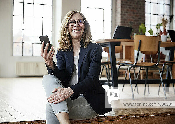 Happy businesswoman with smart phone sitting on hardwood floor