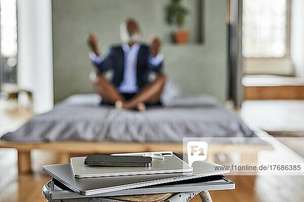Gadgets on table with businessman meditating on bed at home