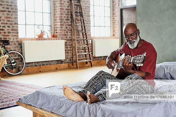 Man with eyeglasses playing guitar on bed at home