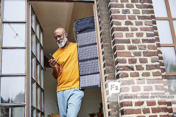 Bald man using smart phone standing with solar panel by window at home