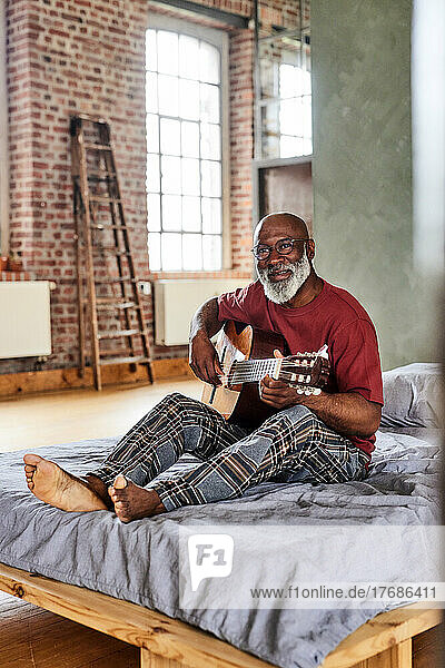 Mature man with eyeglasses playing guitar on bed at home