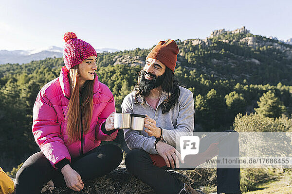 Smiling man and woman toasting mugs sitting on rock