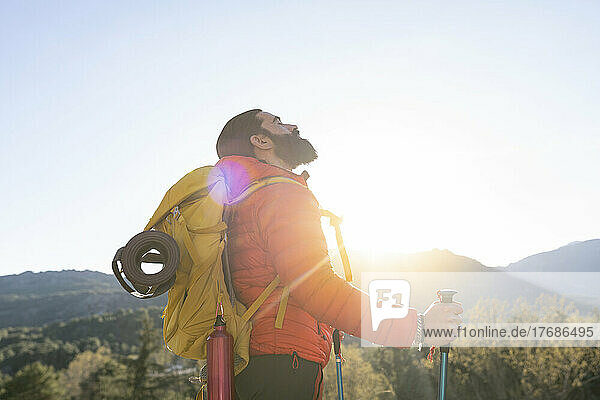 Bearded man wearing backpack standing with hiking poles on sunny day