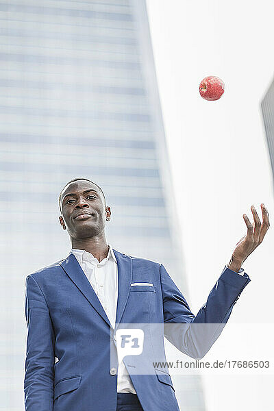 Confident young businessman catching apple in front of modern office building