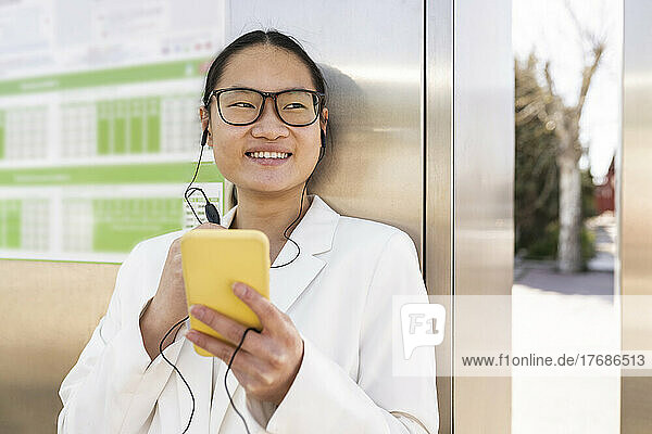 Lächelnde Geschäftsfrau hält Smartphone in der Hand und hört Musik über In-Ear-Kopfhörer
