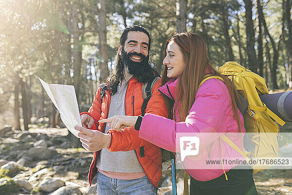Happy bearded man with map looking at girlfriend wearing backpack in forest