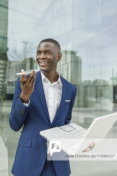 Smiling businessman holding laptop talking on mobile phone through speaker