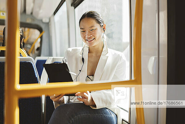 Smiling businesswoman holding tablet PC listening music through in-ear headphones in tram