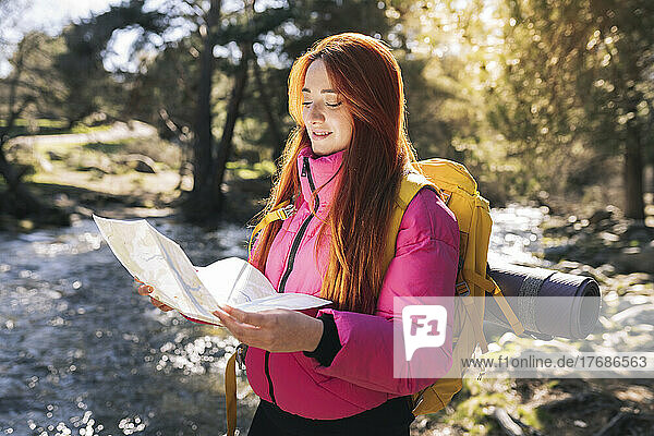 Smiling woman wearing backpack reading map standing in forest