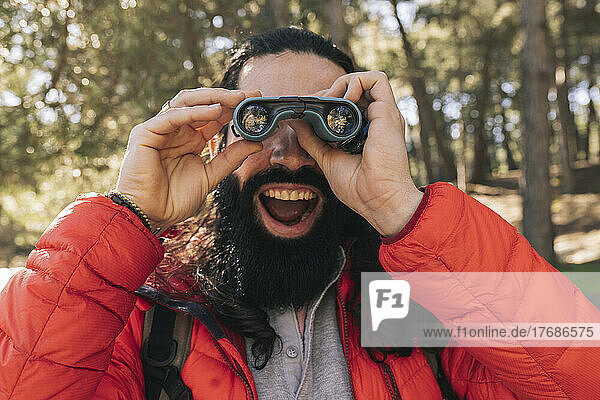 Cheerful bearded man with mouth open looking through binoculars in forest