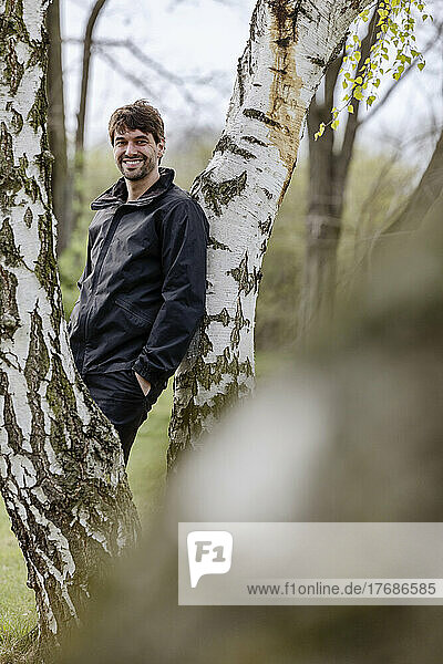 Happy man with hand in pocket leaning on tree
