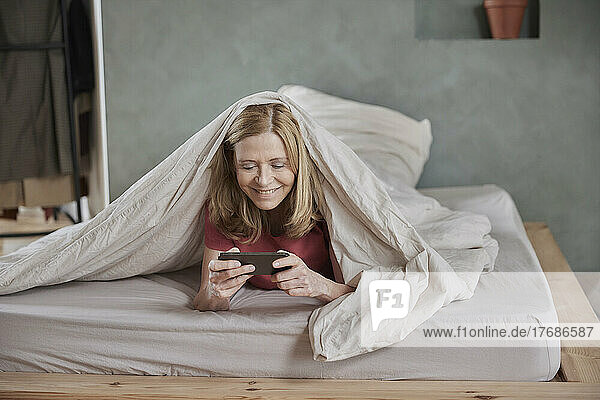 Smiling woman using smart phone lying under blanket on bed at home
