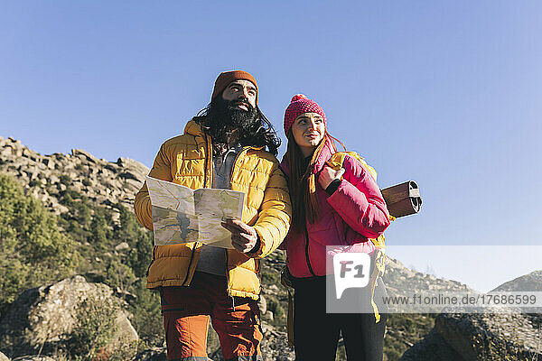 Bearded man with map standing by girlfriend wearing backpack on sunny day