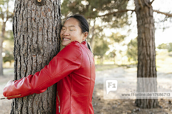 Happy young woman wearing red jacket embracing tree in forest