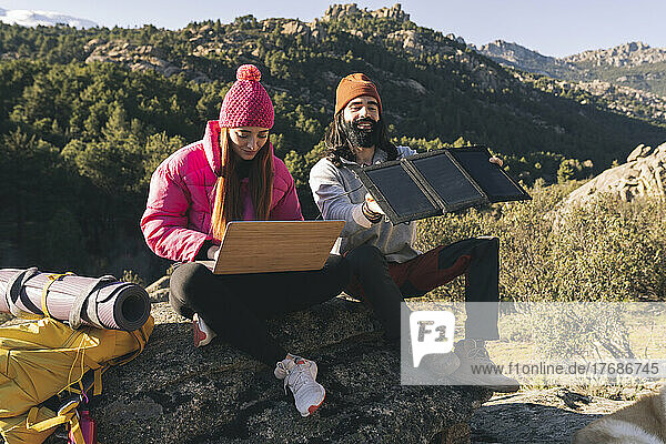 Young woman using laptop sitting by man adjusting solar panel on sunny day