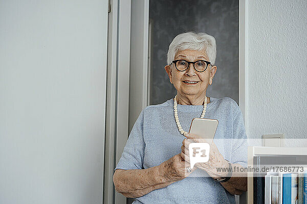 Lächelnde ältere Frau mit Mobiltelefon steht zu Hause
