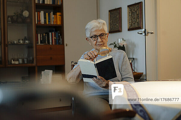 Ältere Frau liest zu Hause Buch mit Lupe