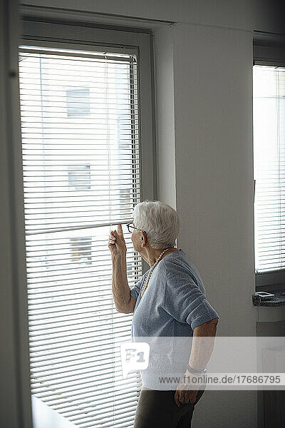 Senior woman looking outside through window blinds at home