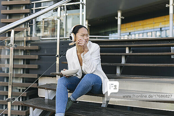 Smiling businesswoman holding bowl listening music through wireless headphones sitting on steps