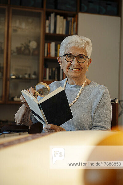 Glückliche ältere Frau mit Buch und Lupe zu Hause