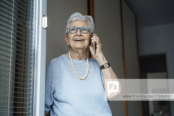 Happy senior woman talking on smart phone standing at apartment doorway