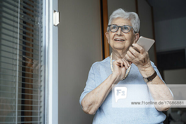 Smiling senior woman with smart phone standing by door of apartment