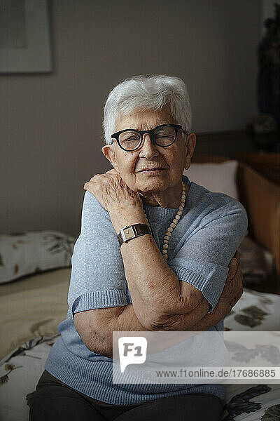 Senior woman with eyes closed hugging herself sitting on bed at home
