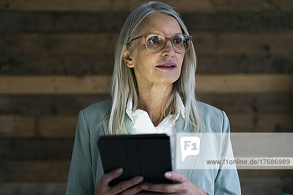 Contemplative businesswoman holding tablet PC in front of wall
