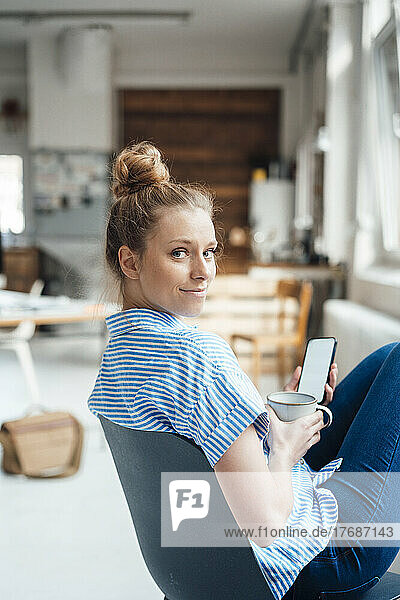 Businesswoman with drink and smart phone sitting on chair at home