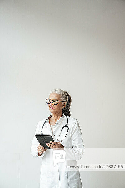 Senior doctor with tablet PC standing against white background