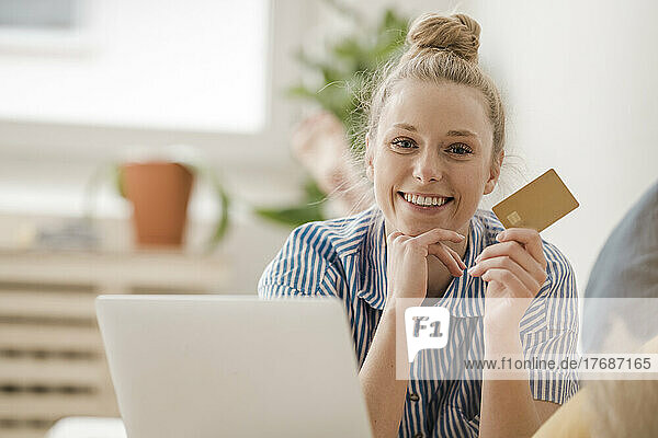 Smiling businesswoman with hand on chin showing credit card at home